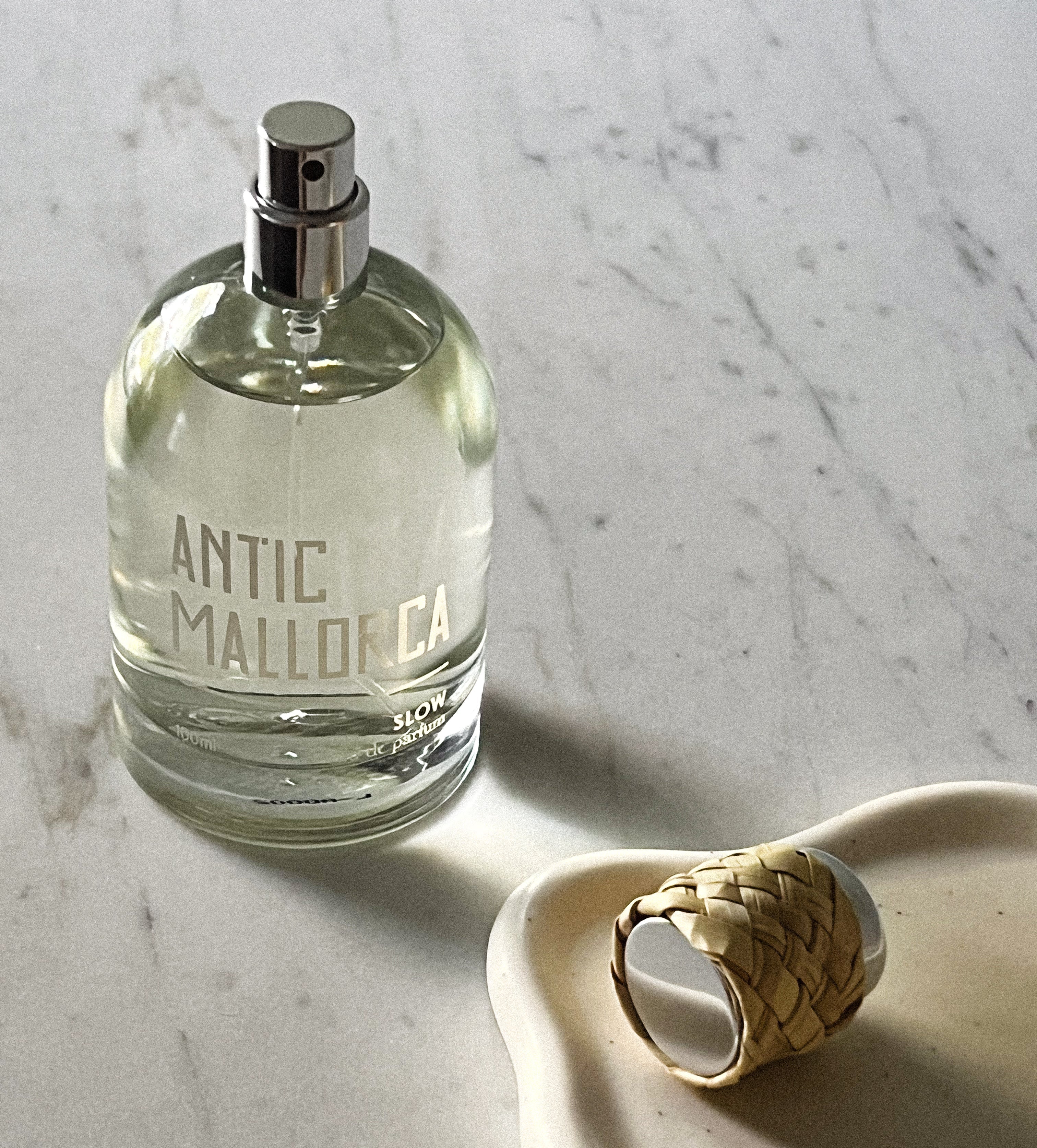 Perfume bottle labeled 'Antic Mallorca' on a marble surface with a woven cap beside it.